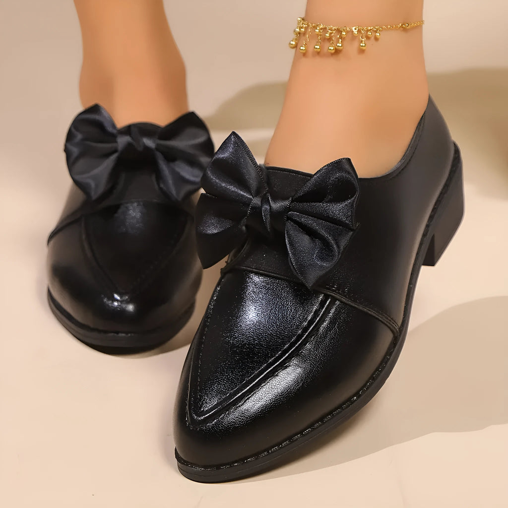 Women's black leather-look flat shoes with a decorative bow on the pointed toe.