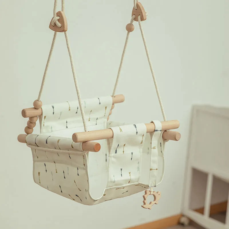 Natural beige cotton canvas baby swing chair with wooden seat and decorative tassel pattern, hanging indoors.