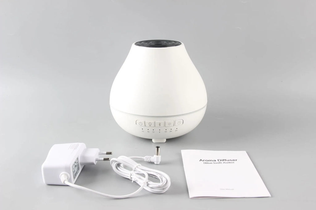 Round white Bluetooth aroma diffuser and humidifier with a colour-changing night light on a table.
