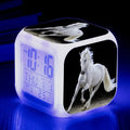 Small square LED digital alarm clock with colour-changing display on a bedside table.