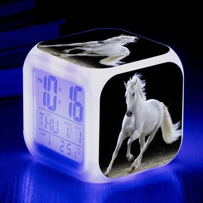 Small square LED digital alarm clock with colour-changing display on a bedside table.