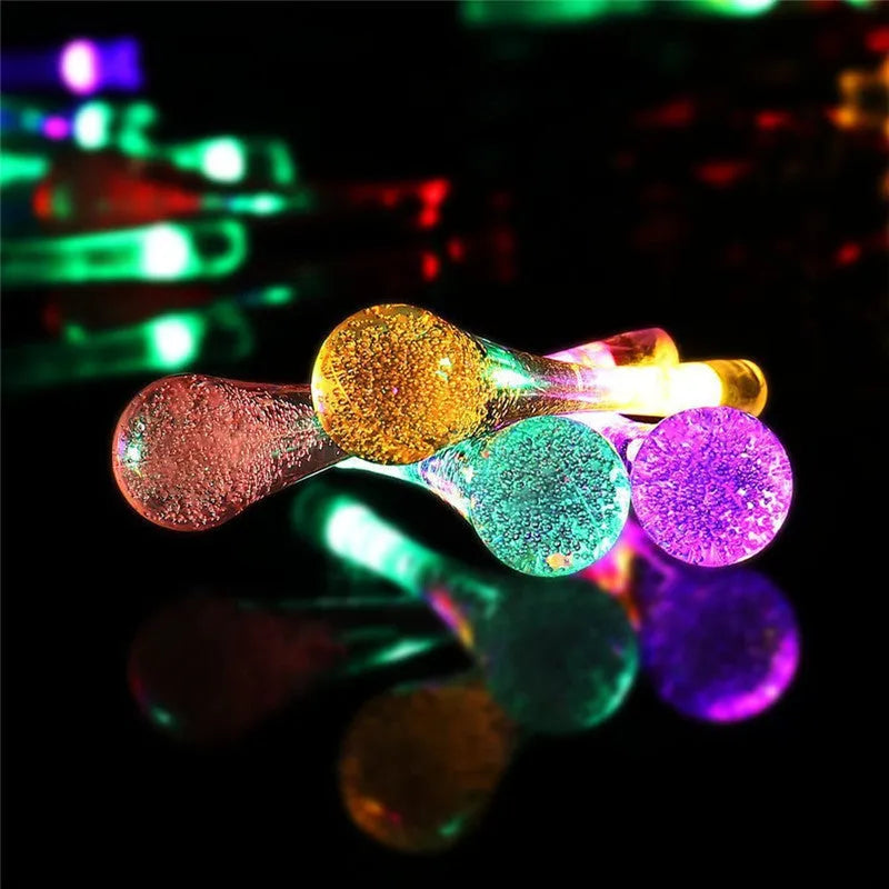 Water drop shaped solar LED fairy lights hanging outdoors with solar panel and ground spike.
