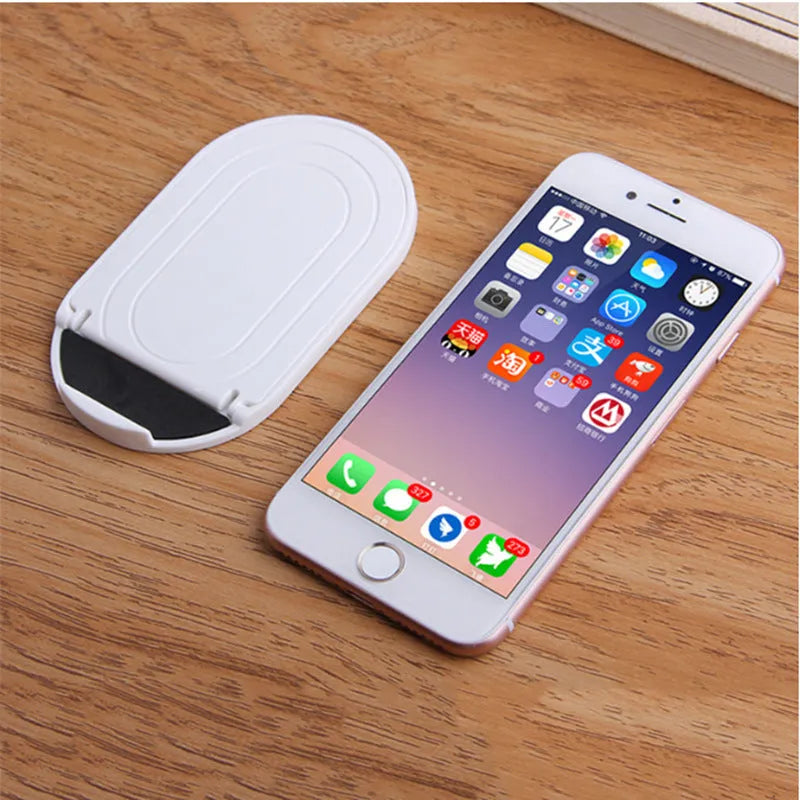 Plastic lazy bracket mobile phone stand on a desk, shown in assorted colours including black, blue, and pink.