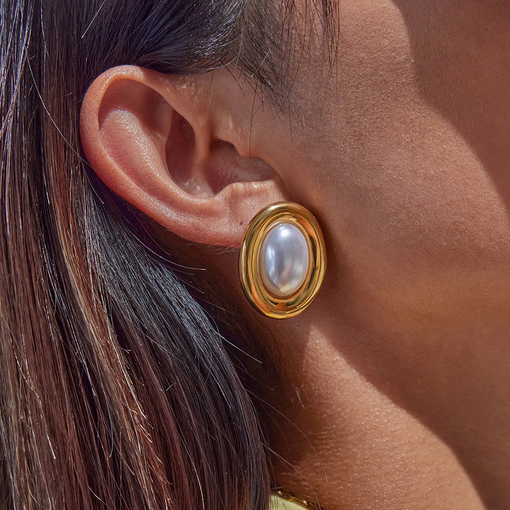 Close-up of a pair of round stainless steel stud earrings with a vintage-style pearl detail on a neutral background.