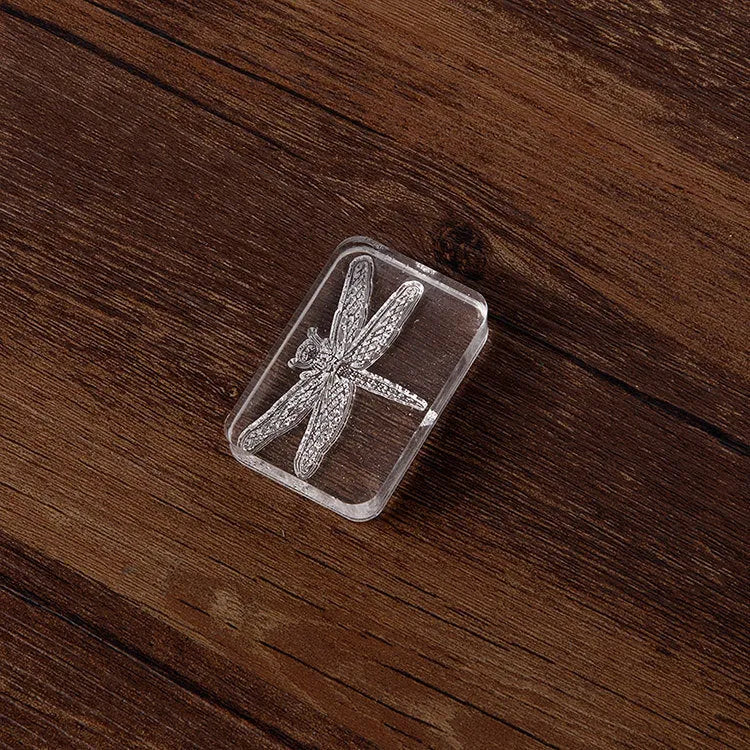 Transparent acrylic hand seal with clear silicone stamp on a white card, showing dragonfly or fern engraving.