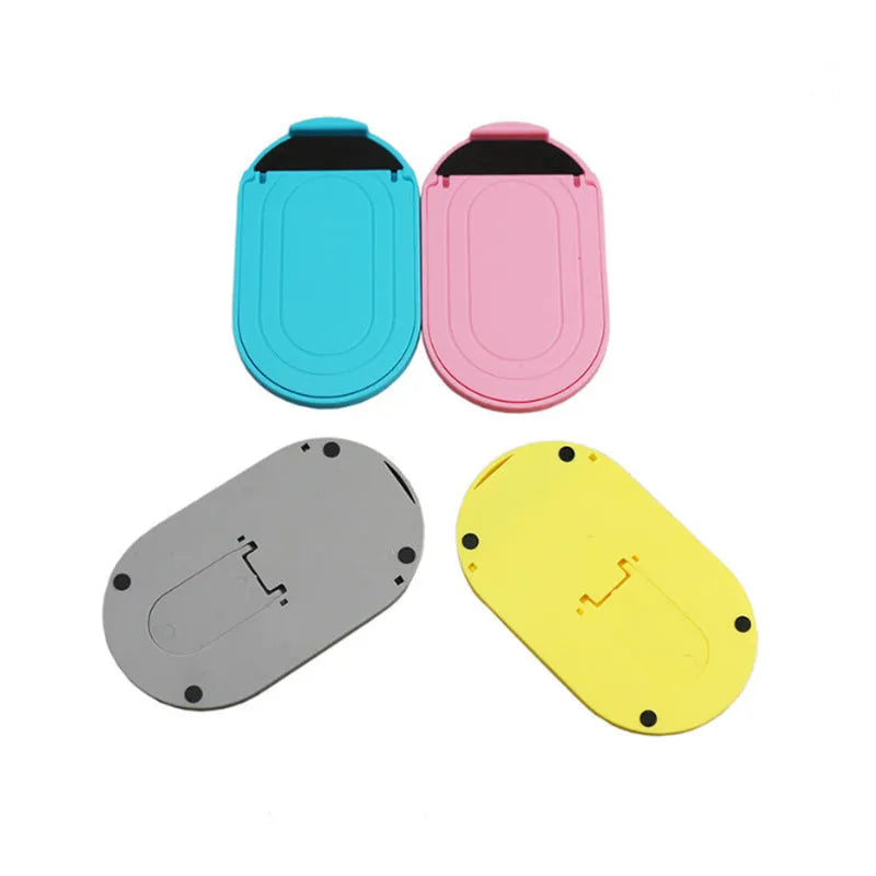 Plastic lazy bracket mobile phone stand on a desk, shown in assorted colours including black, blue, and pink.
