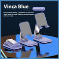 Folding portable desktop mobile phone stand in black, white, blue, or pink on a wooden desk.
