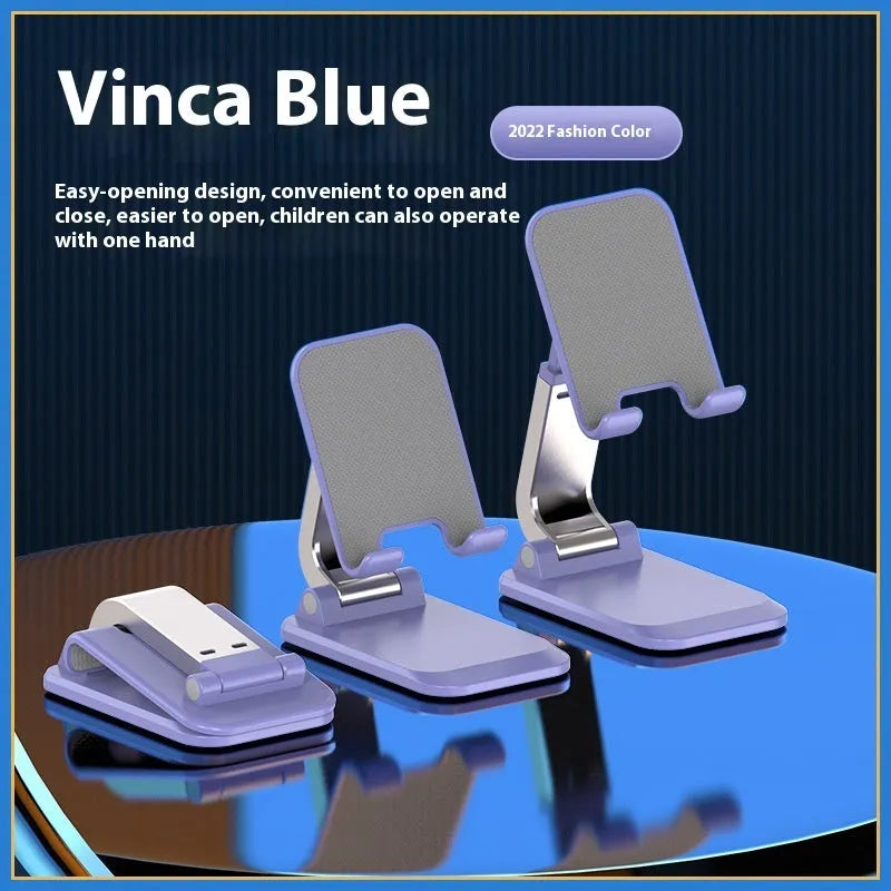 Folding portable desktop mobile phone stand in black, white, blue, or pink on a wooden desk.