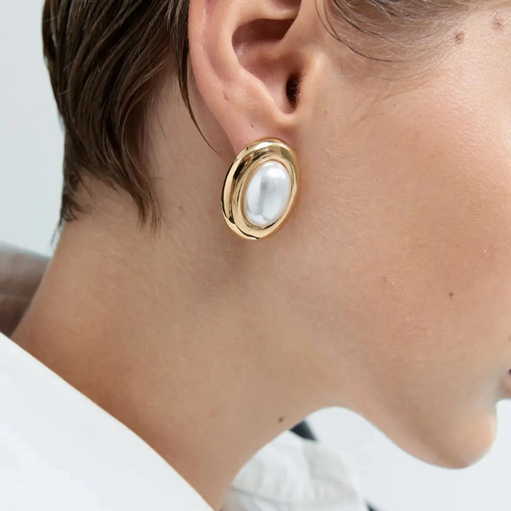 Close-up of a pair of round stainless steel stud earrings with a vintage-style pearl detail on a neutral background.