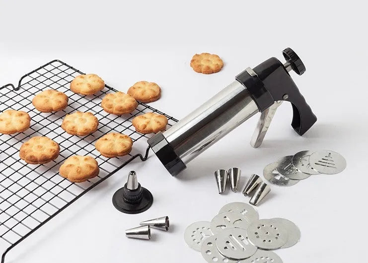 Stainless steel biscuit press gun with assortment of cookie moulds and piping nozzles on a baking sheet.