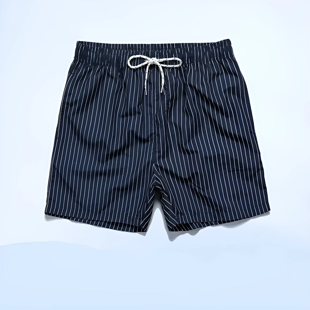 Man wearing navy blue and white striped straight-leg polyester beach shorts on shore.