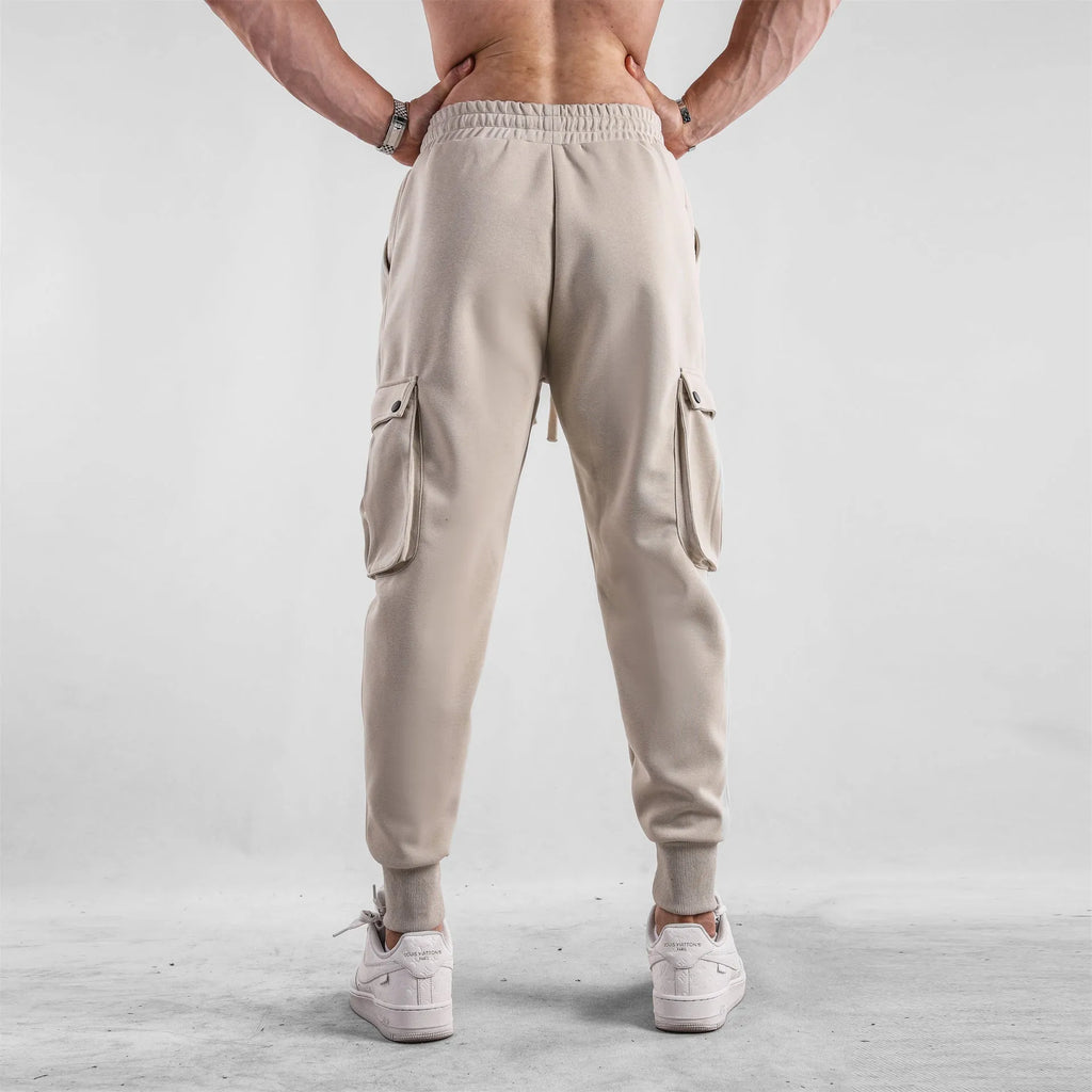 Men's loose beige cotton trousers with an ankle tie and multiple pockets on a casual model.