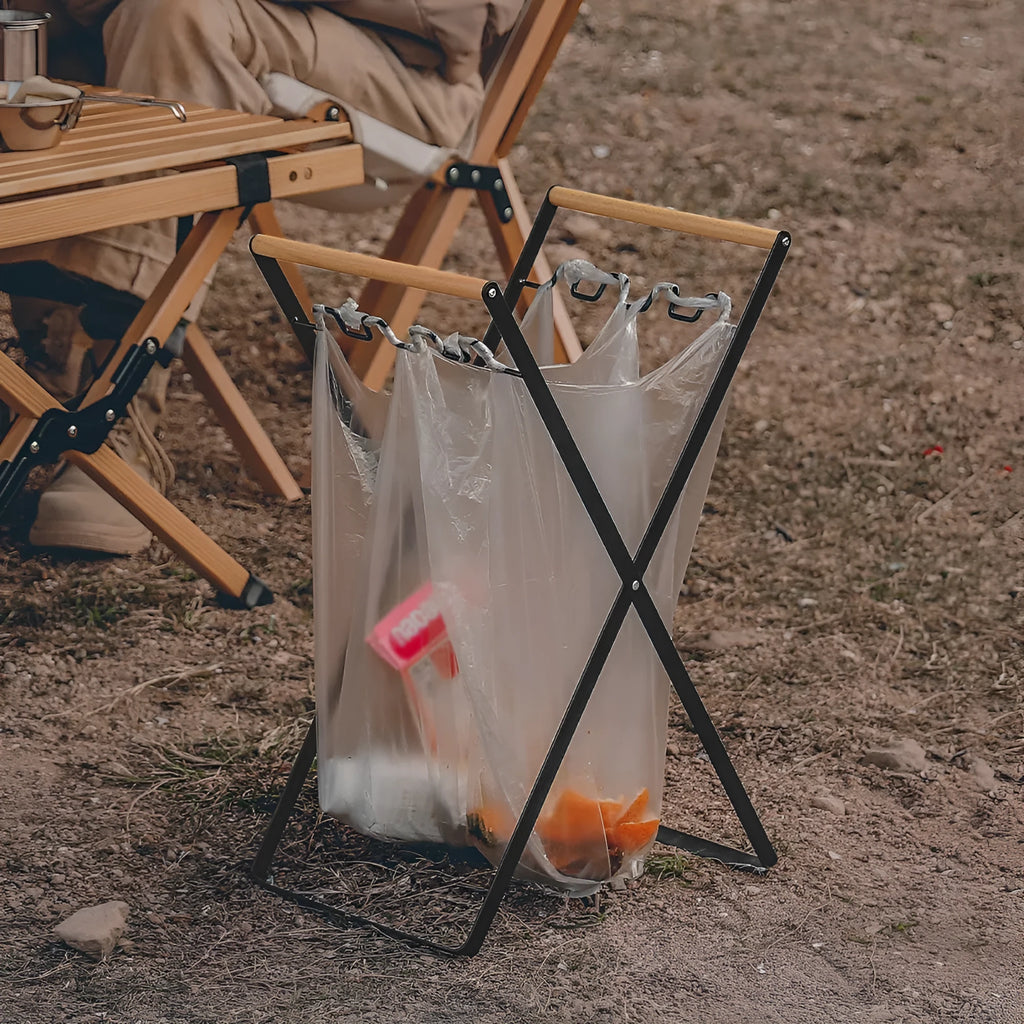 Black iron folding bin rack with wooden handles holding two rubbish bags in an outdoor setting.