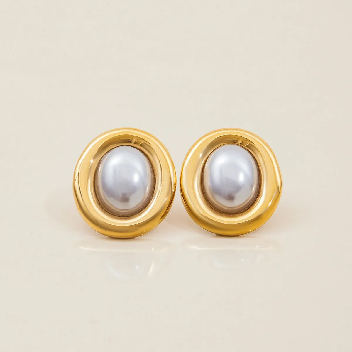 Close-up of a pair of round stainless steel stud earrings with a vintage-style pearl detail on a neutral background.
