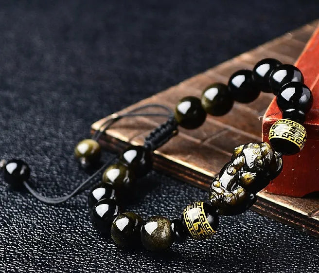 Close-up of a black obsidian bead bracelet with a detailed silver Pi Xiu charm on a neutral background.