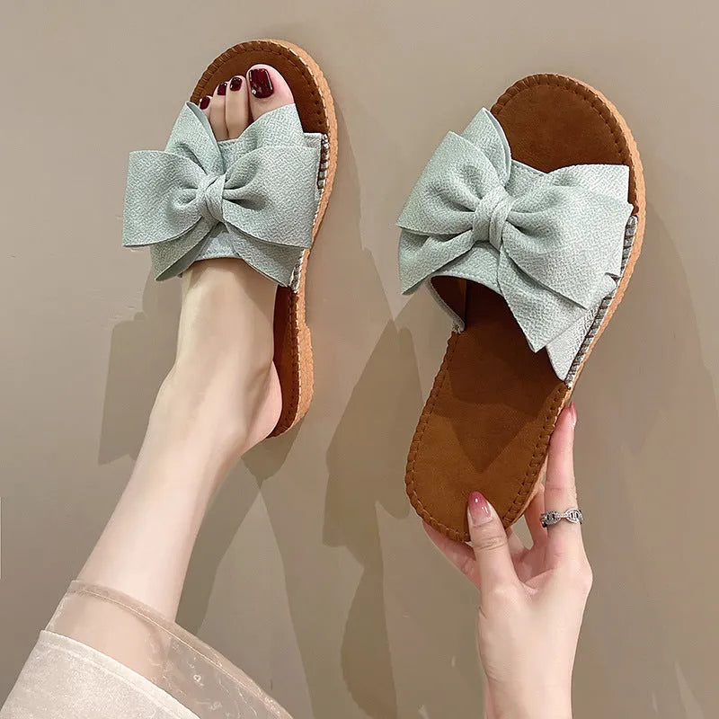 Women's beige slip-on sandals with a suede cloth upper, soft rubber sole, and decorative bow.
